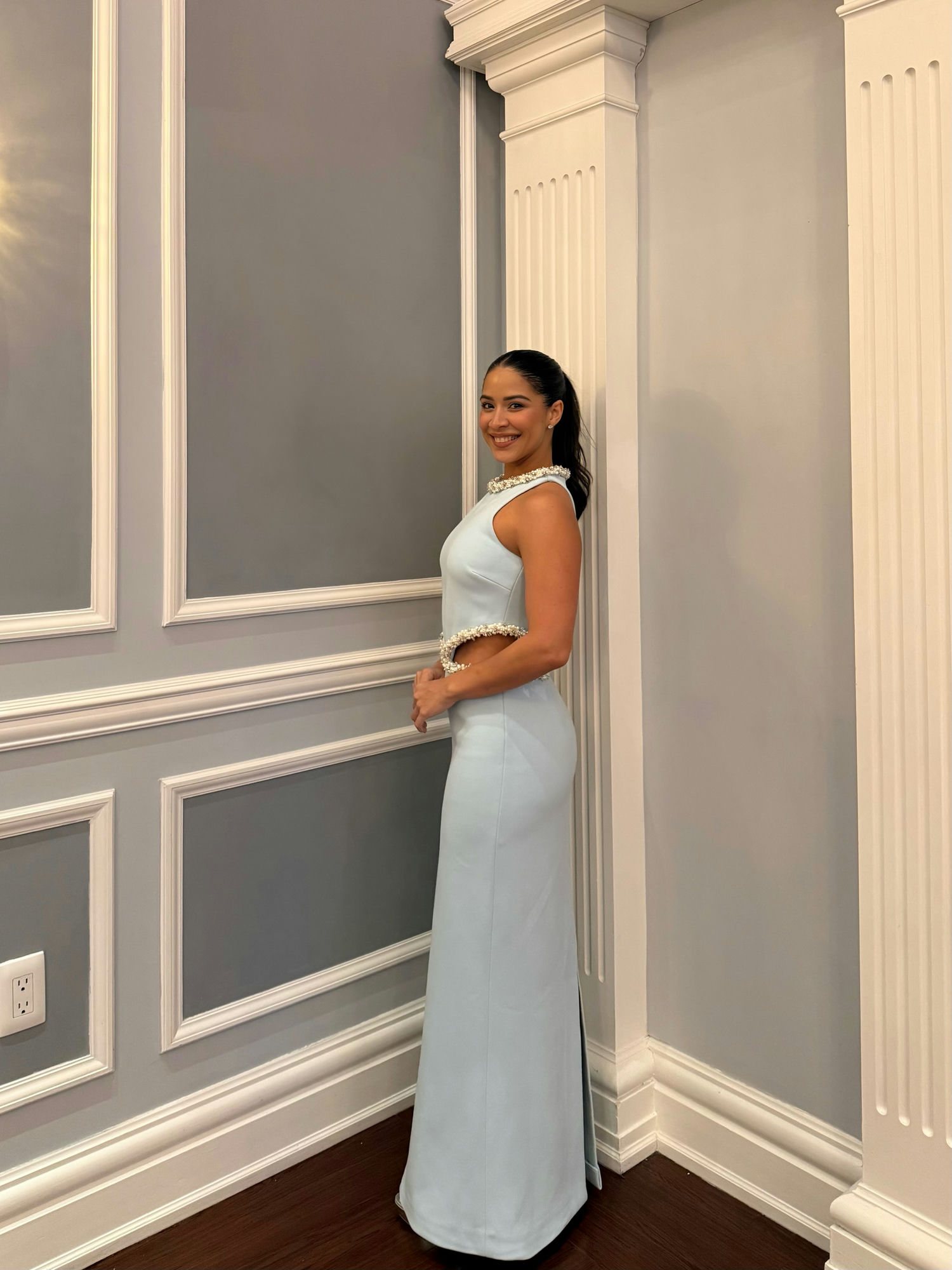 Mujer con un vestido largo azul claro de dos piezas frente a un muro gris con paneles